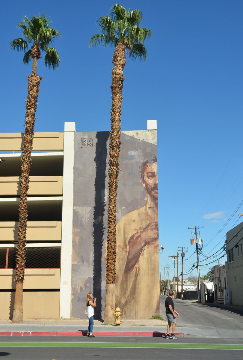 a large mural on the corner of a 4 storey parking garage, a man from the waist up who is looking down on the sidewalk - viewed from straight on so only one half of the mural, and one half of the man, is visible