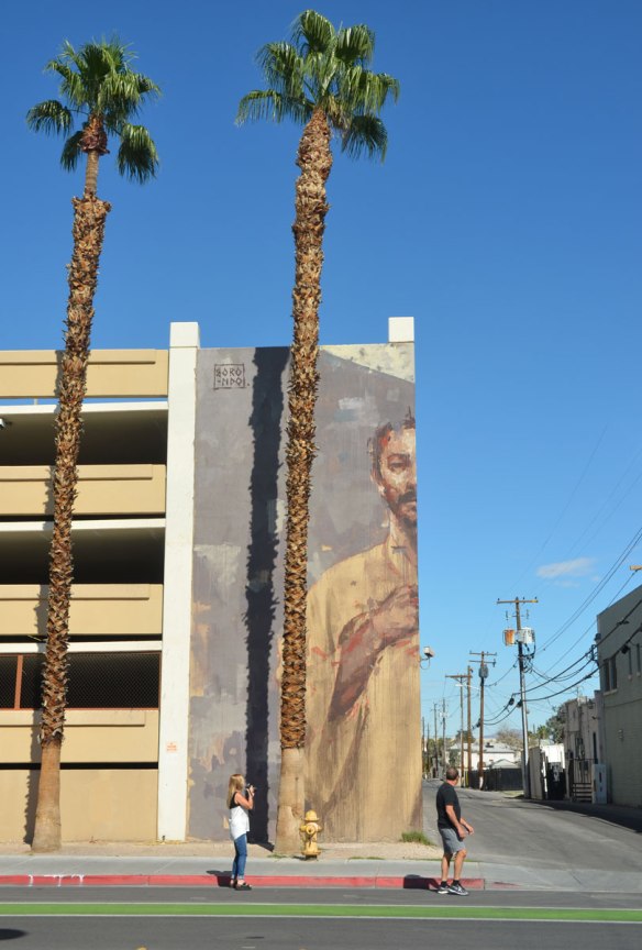 a large mural on the corner of a 4 storey parking garage, a man from the waist up who is looking down on the sidewalk - viewed from straight on so only one half of the mural, and one half of the man, is visible
