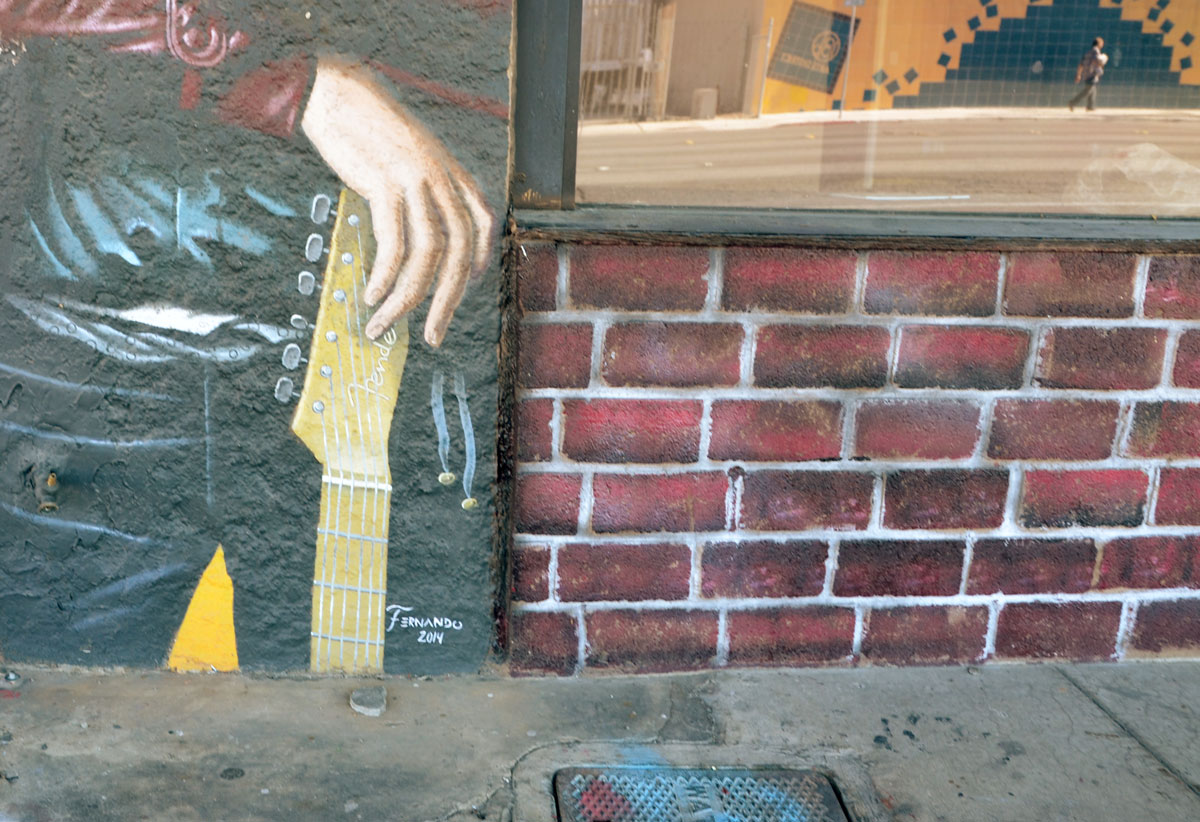 signature at the bottom of a portrait of a guitar player, a hand holding a fender guitar is in the picture along with part of the brick wall of the building. 
