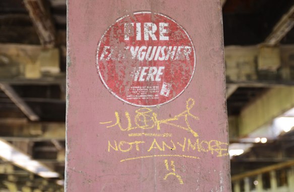 pink concrete post with an old painting on it, red circle with white words that says fire extinguisher here. Someone has written in yellow below it, not anymore. 