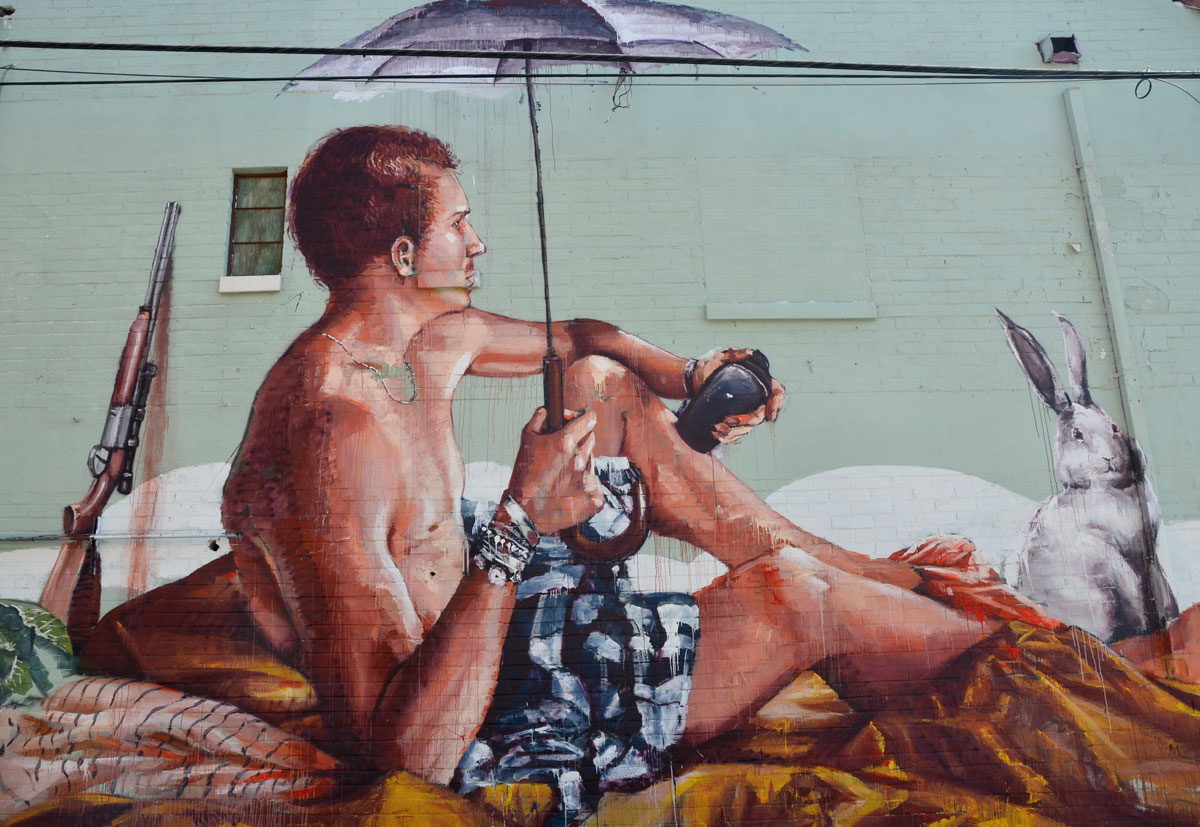 mural of a man holding an umbrella as he sits on the ground. A rifle is behind him. He is holding a small black case in one hand. 