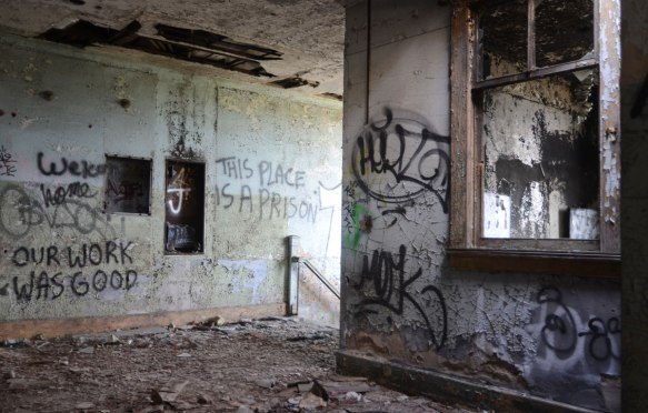 Words and other graffiti written on grey walls of an abandoned train station in Buffalo, one set of words says This place is a prison. Another set of words says Our work was good 
