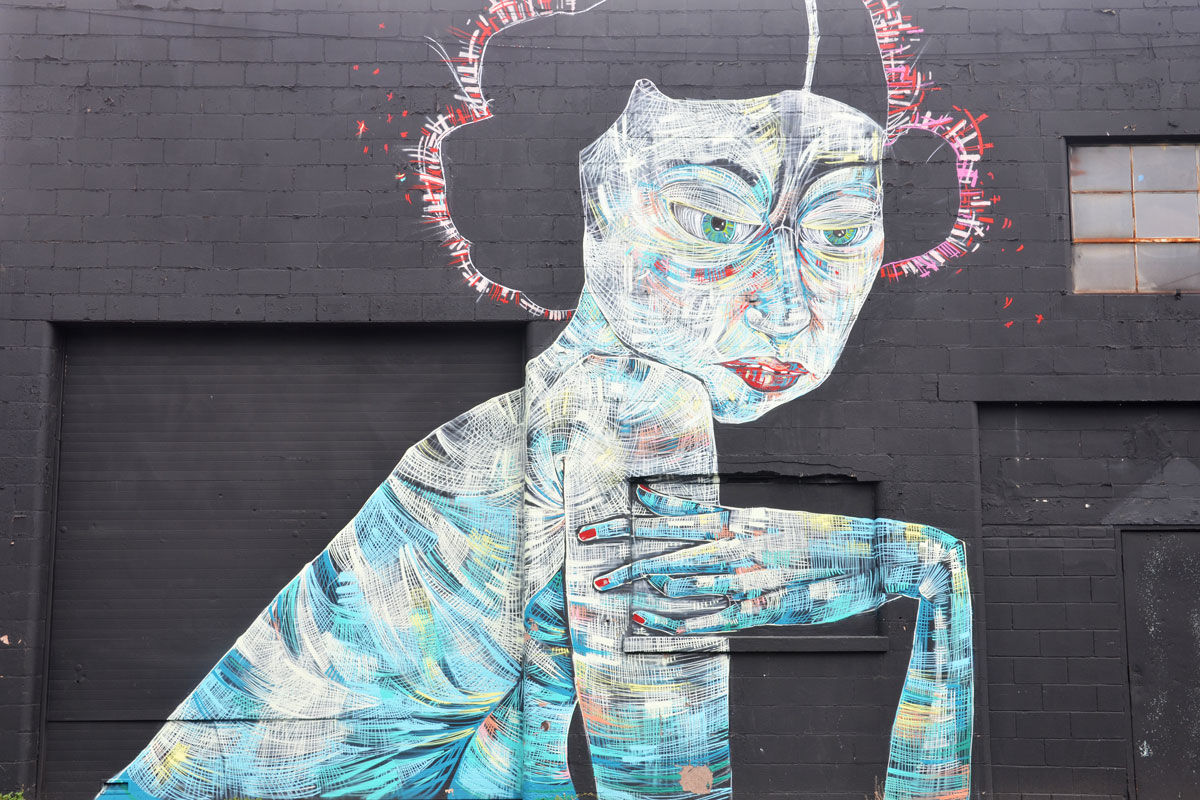 mural on the side of a black brick building, of a woman done in wide strokes of different blues, yellows and white. Upper body, mostly in profile, with her elbow bent and her hand turned towards her arm. 