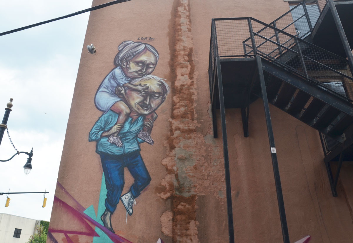 mural on the back of a red brick building, beside a metal exterior staircase, of a man carrying a woman on his back. Above the picture are the words I Got You