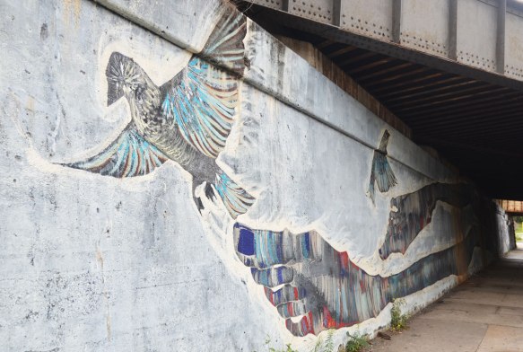 part of a long mural under a railway bridge, the end of the mural with a person's feet and large toes, with two birds also in the picture. 
