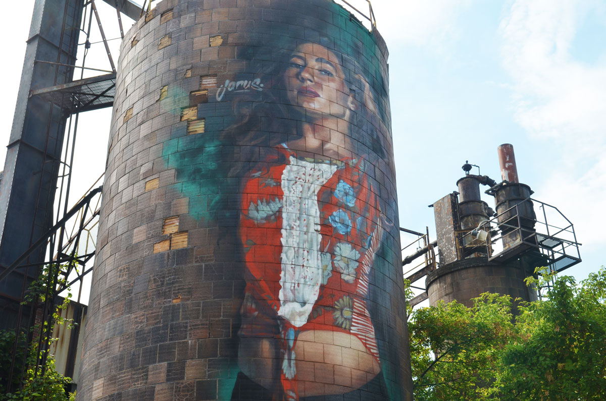 A mural by Jarus of a woman on a water tower (or silo shaped storage tower). 