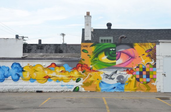 A mural on the side of a building in Rochester New York showing a large eye that is half green and half pink. Lots of swirls of colour as well. Also a small section is painted as a grid of different colours. 