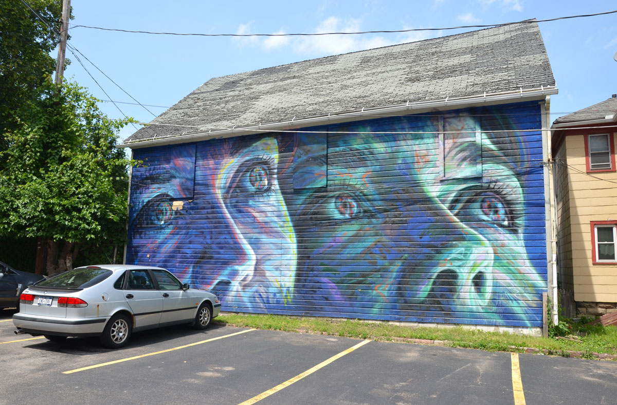 Wall Therapy mural in Rochester, on the side of a building, two large bluish faces looking up towards the sky. A car is parked in front of it. 