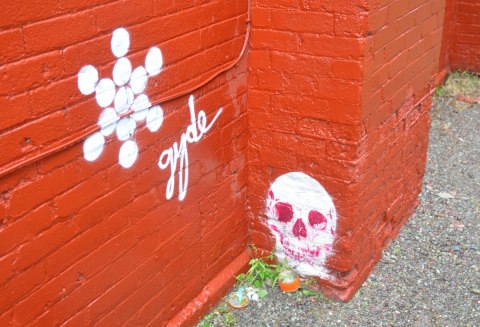 brick wall painted orange. A skull graffiti stencil is on the wall, just above ground level. A snowflake shaped graffiti in white next to it with the word gyde written in cursive