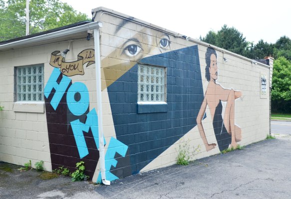 mural on the corner of a building, The word home is written in large blue letters around the corner, and on one side of the building is a pair of eyes over a real window, and a woman sitting beside the window. It looks like she is sitting on the ground, one knee up and she is turned from the waist to look back at the viewer. 