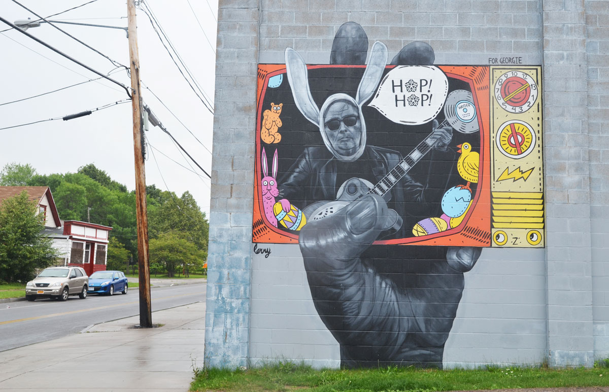Wall Therapy mural in Rochester, on the side of a building, a very large hand is hold a TV screen. A man with big bunny ears seems to be coming out of the screen along with a word bubble that says "hop hop"