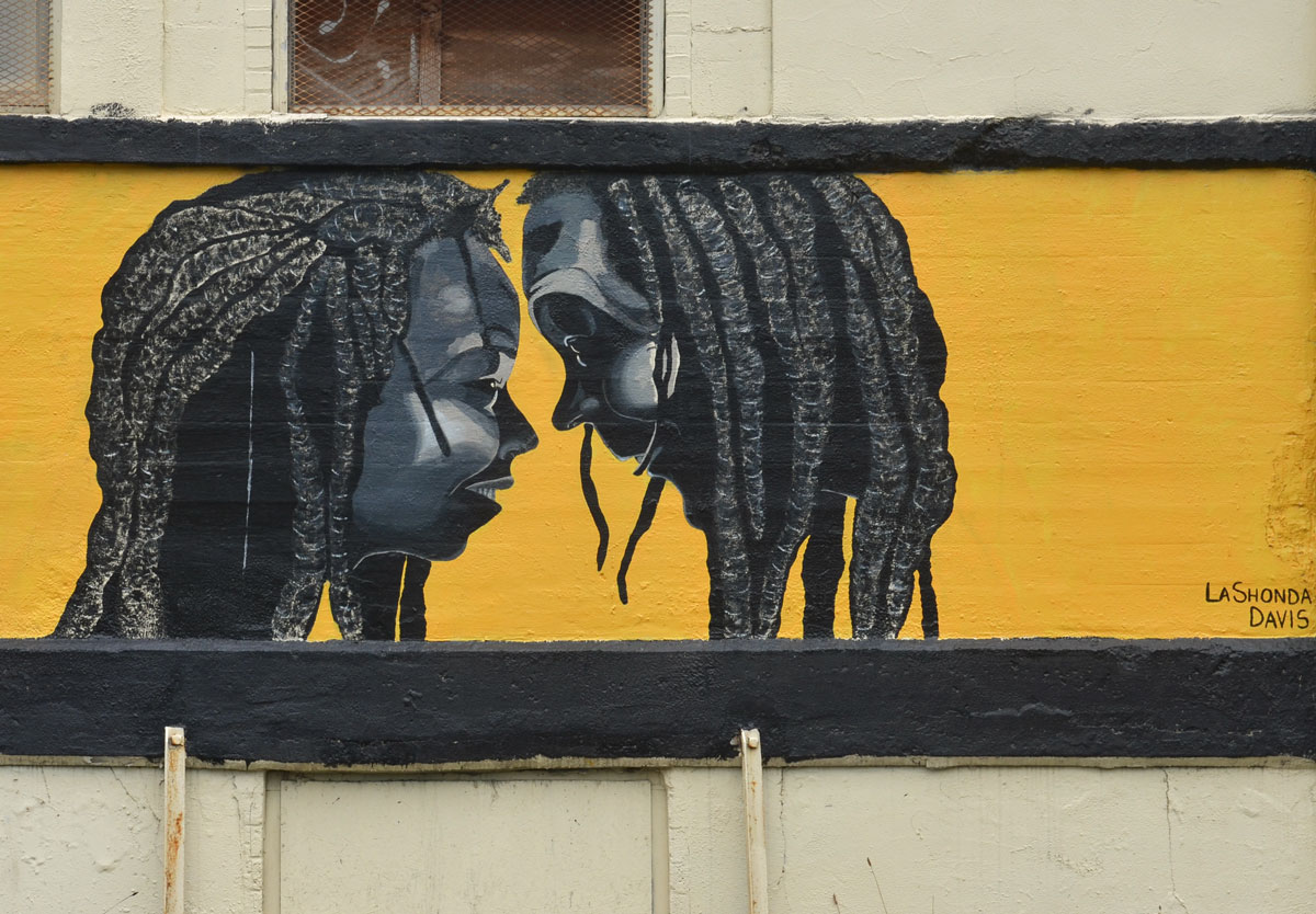 A mural by LanShonda Davis, yellow background, two faces in profile, both black tieh dreadlocks, one man, one woman, they are face to face and their noses are almost touching. 