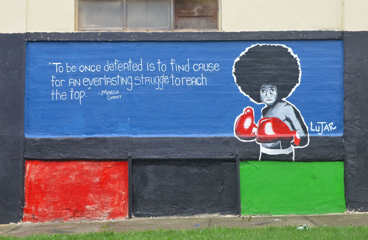 A mural on the side of a building. Blue background. A young man with an Afro hair style is wearing red boxing gloves. The words on the mural say " To be once defeated is to find cause for an everlasting struggle to reach the top" It is a quote attributed to Marcus Garvey. The mural is signed by Lujar. 