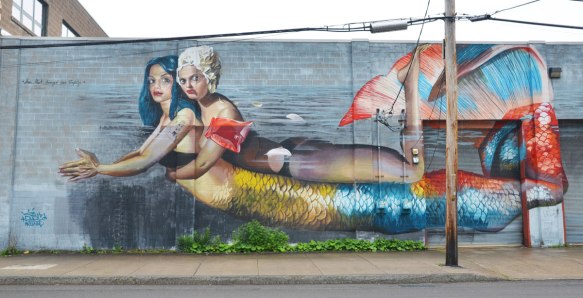 A large mural of a mermaid swimming in the water with another woman on her back - the other woman is wearing a white bathing cap and orange arm floaties. 