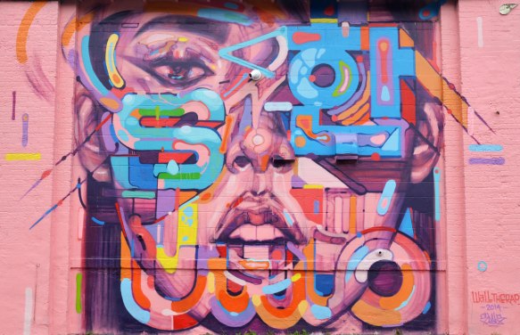 Mural of a large pink and purple face with letters in different colours painted on top of parts of it. One eye, nose and open mouth are visible.