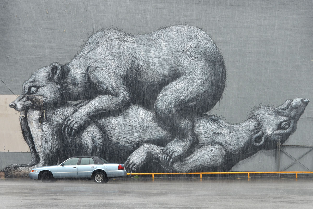 Black and white and grey mural, very large, on the side of a building by a parking lot by ROA featuring 2 bears, one on top of the other. A car is parked in front of the mural. It is raining. 