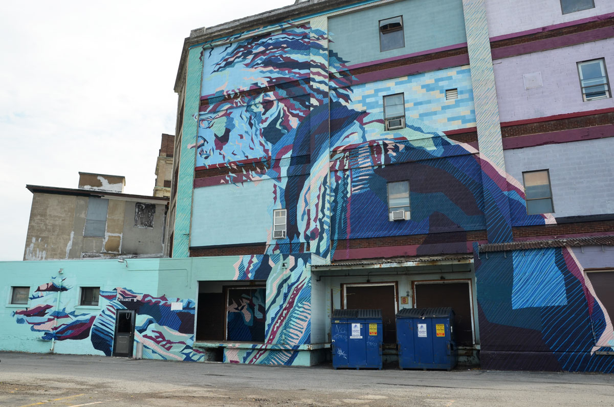 A large four storey mural of a man, upper body, in profile, in blues and purples. His hands are in front of him. 