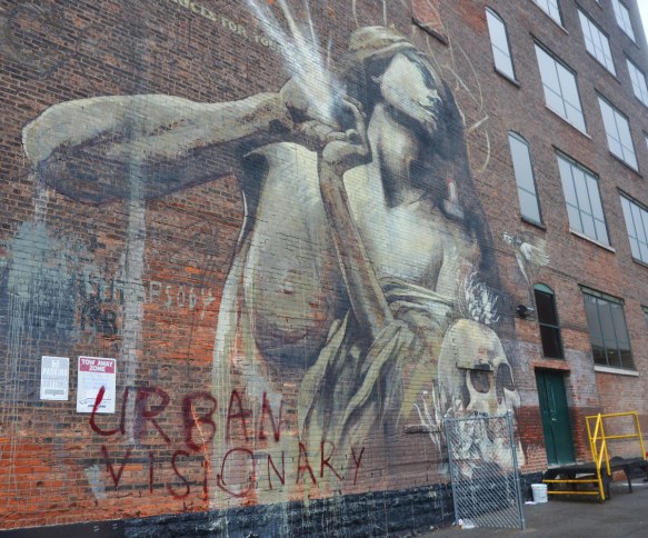 mural of a woman looking upward with a light radiating from her hands, a skull is below and beside her. 
