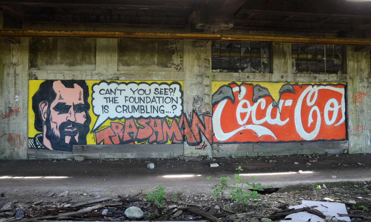 graffiti painting - a man with the words Can't you see? The foundation is crumbling. Trashman is written in large red letters, also a mock coca-cola logo 