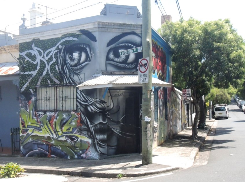 A house on a corner that has been covered with a mural of a woman's face in black, white and grey. Her nose and mouth are on the first storey, her eyes are on the upper storey
