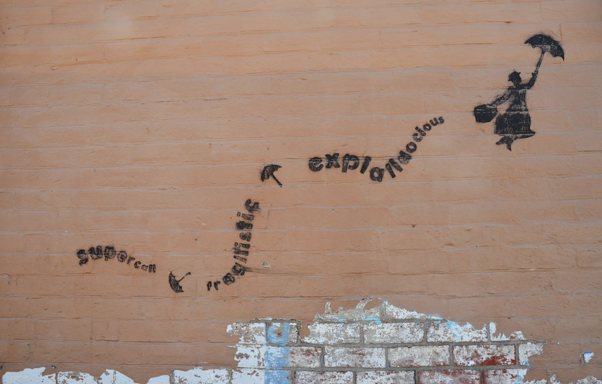 black stencil on a light brown wall, Mary Poppins floating upwards with her open umbrella. Her trail is the word supercalifragiliciousexpialidocius. 