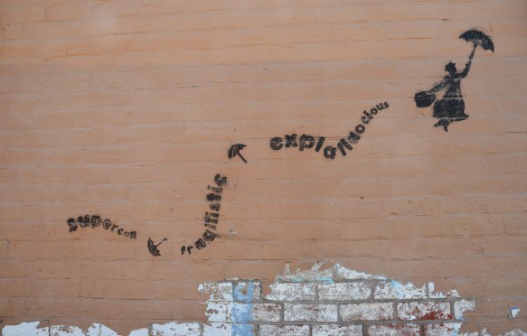 black stencil on a light brown wall, Mary Poppins floating upwards with her open umbrella. Her trail is the word supercalifragiliciousexpialidocius. 
