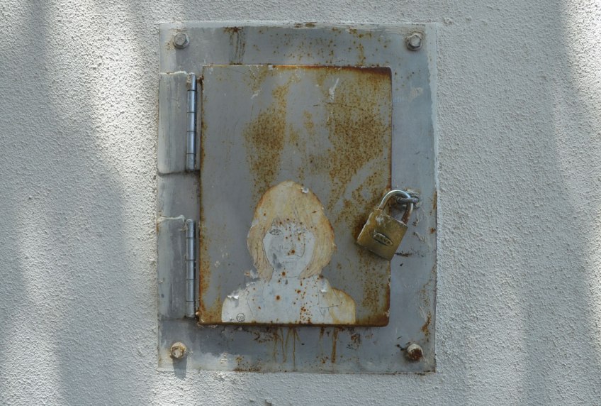 small paper wheatpaste of a blond woman on the door of a small metal box, with a padlock. The paper is peeling a bit and the rust from the box is also on the graffiti