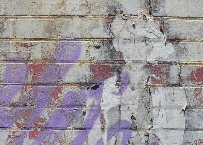 Paper wheatpaste of a woman that is badly faded and peeled off. She is beside some purple spray paint.