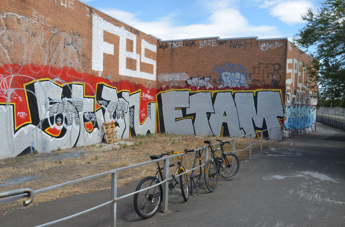 two bikes parked against a metal railing beside a bike path. on the other side of the railing is a two storey L shaped brick building with the lower level covered with two large tags, one says ETAM. FBS is written on the second storey. 