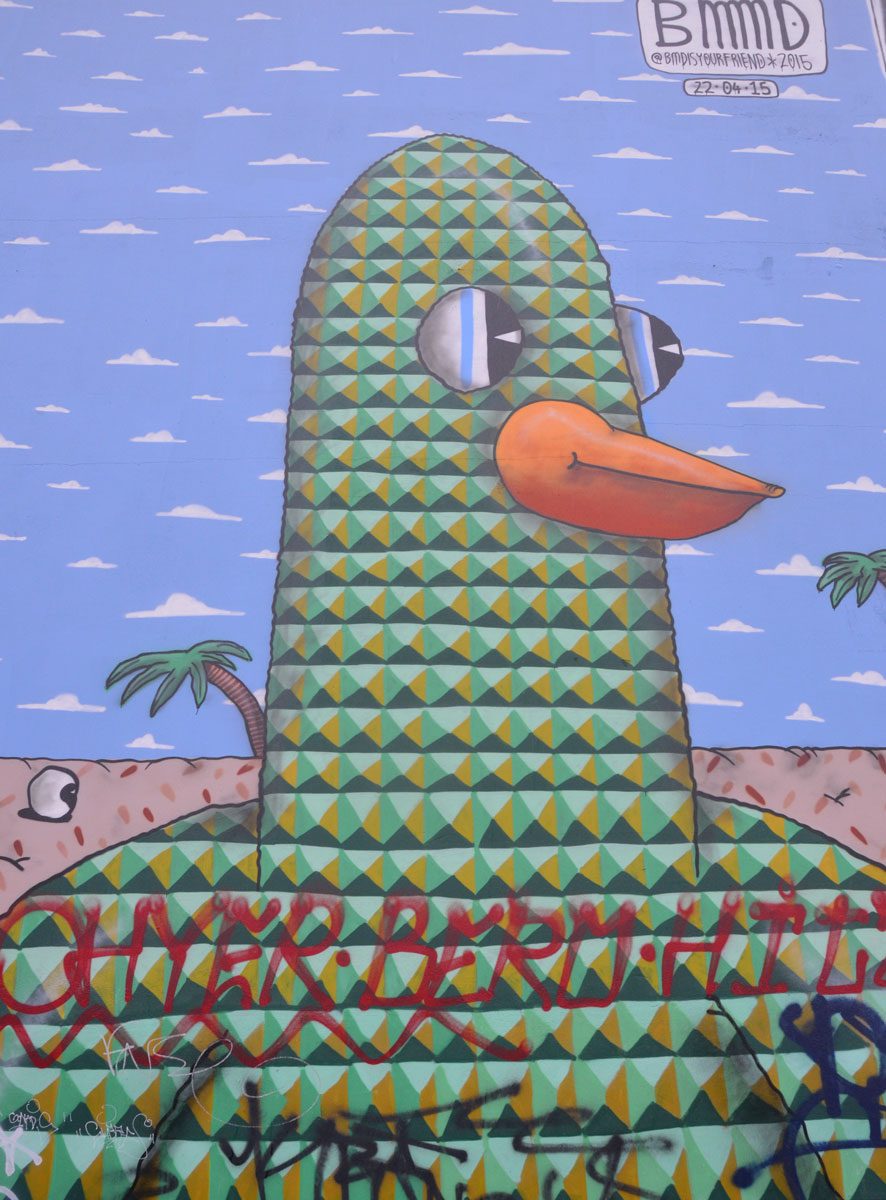 bird head on a mural, drawn with interlocking triangles in green and black, blue sky and palm trees in the background