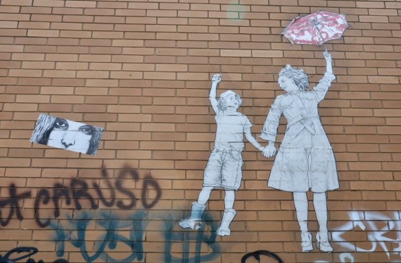 On a brown brick building, a wheatpaste of a mother holding a child's hand and she is holding a red and white parasol in the other. 