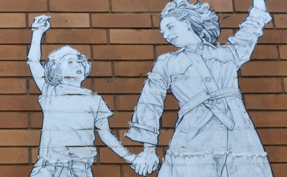 On a brown brick building, a wheatpaste of a mother holding a child's hand and she is holding a red and white parasol in the other. Close up of the upper part of the people