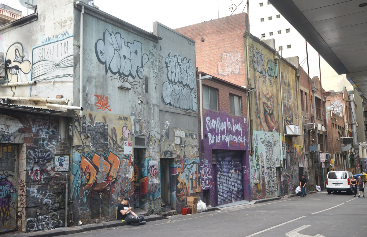 buildings in an alley with graffiti all over them. A man is sitting on the kerb in front of one of the buildings
