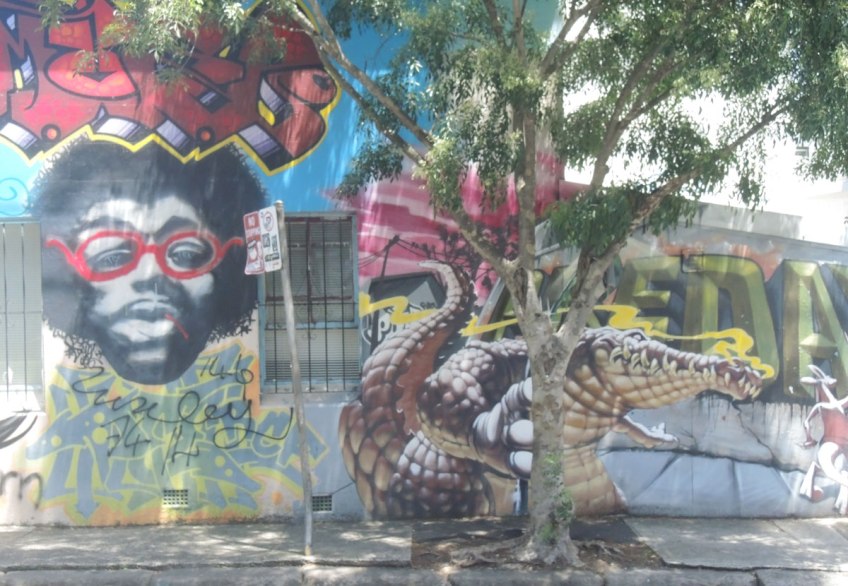 A mural on a wall, includes a black man with beard and mustache and red rimmed glasses, also includes a realistic looking crocodile