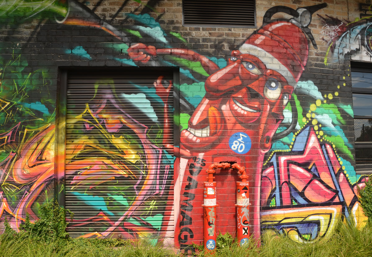 street art of what looks like a tall red fire hydrant with at least two faces, on on the front and one on the side. Arms are hoses, smiling, 
