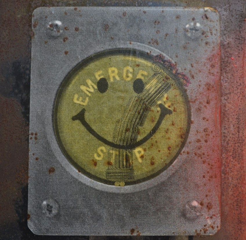 paste-up in a Melbourne lane of a flat piece of metal screwed to a post with 4 screws, one in each corner and a large round emergency stop button in the middle. The round button has been turned into a yellowish happy face
