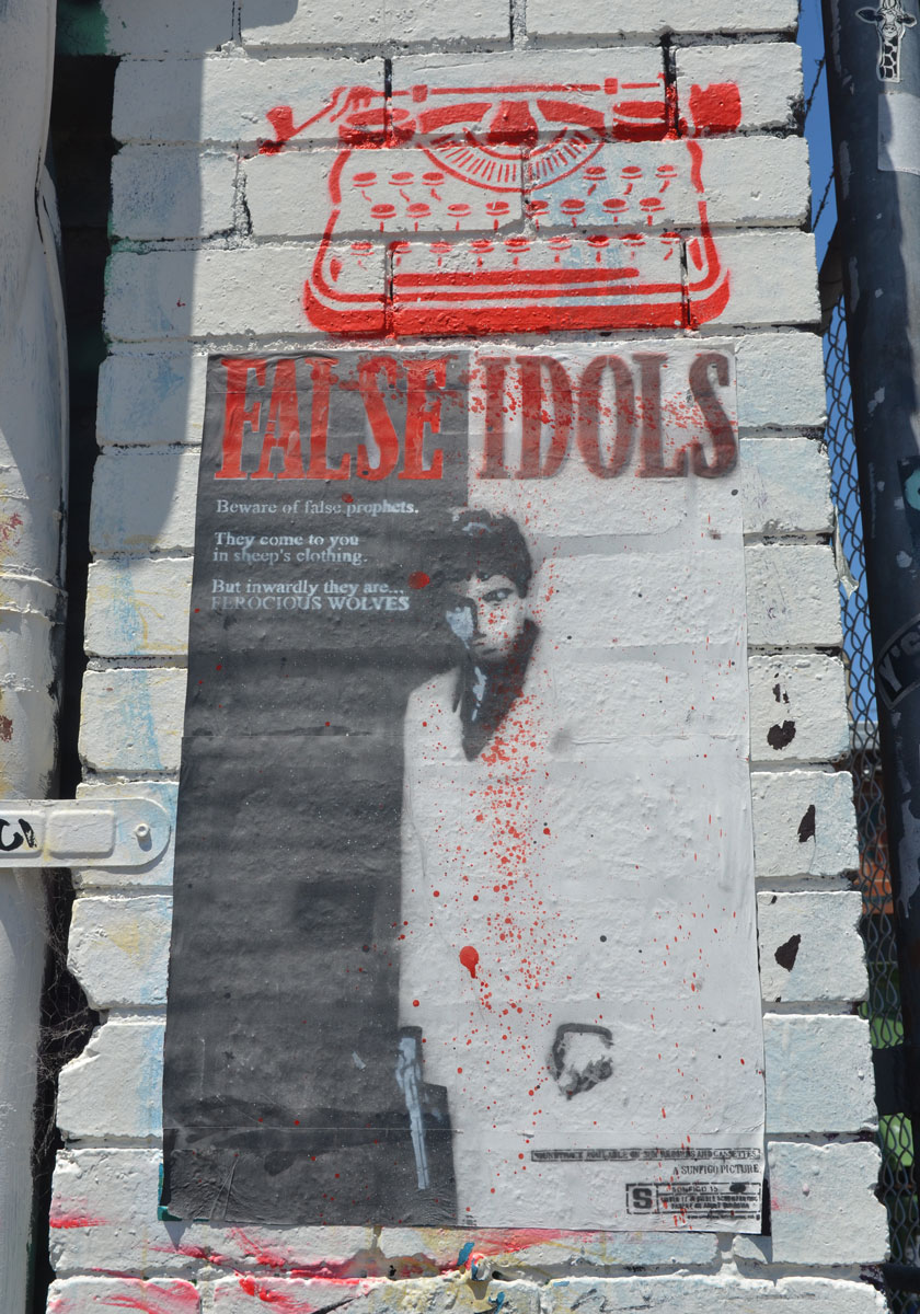 A red typewriter stencil by wrdsmith over a poster for False Idols paste-up on a wall. A picture of a man is on the poster along with the words "Beware of false prophets. They come to you in sheep's clothing. But inwardly they are ferocious wolves " A tiny giraffe head is in the top right corner.