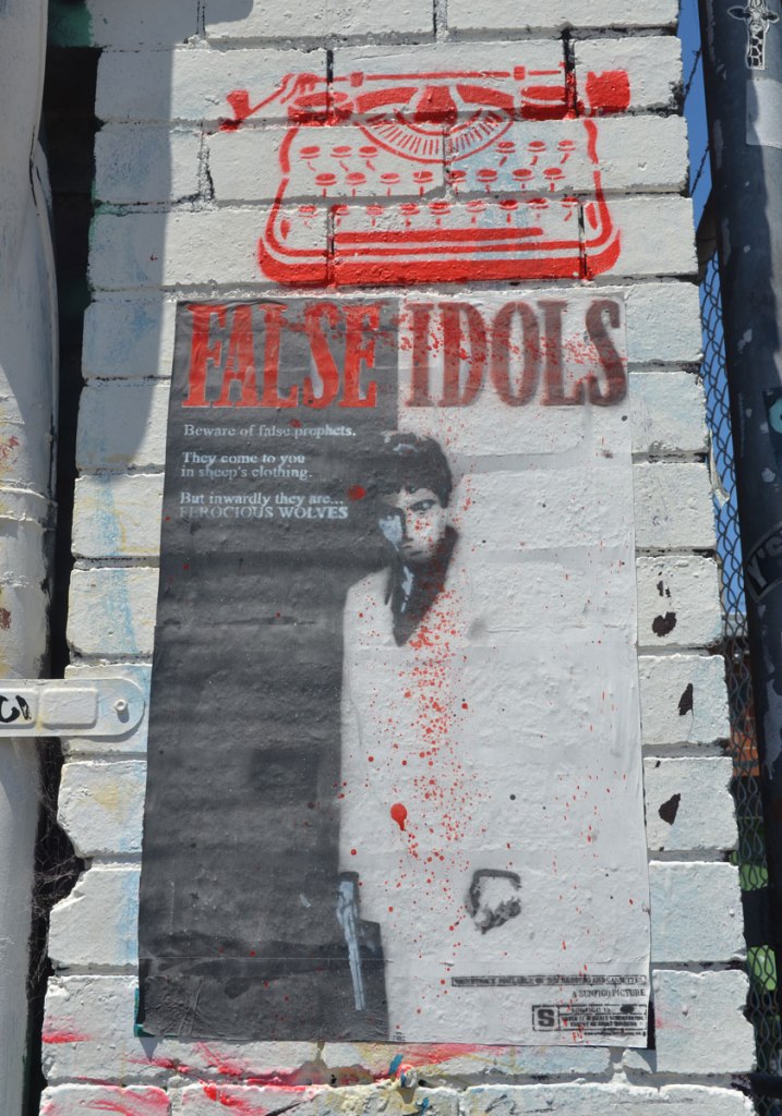 A red typewriter stencil by wrdsmith over a poster for False Idols paste-up on a wall. A picture of a man is on the poster along with the words "Beware of false prophets. They come to you in sheep's clothing. But inwardly they are ferocious wolves