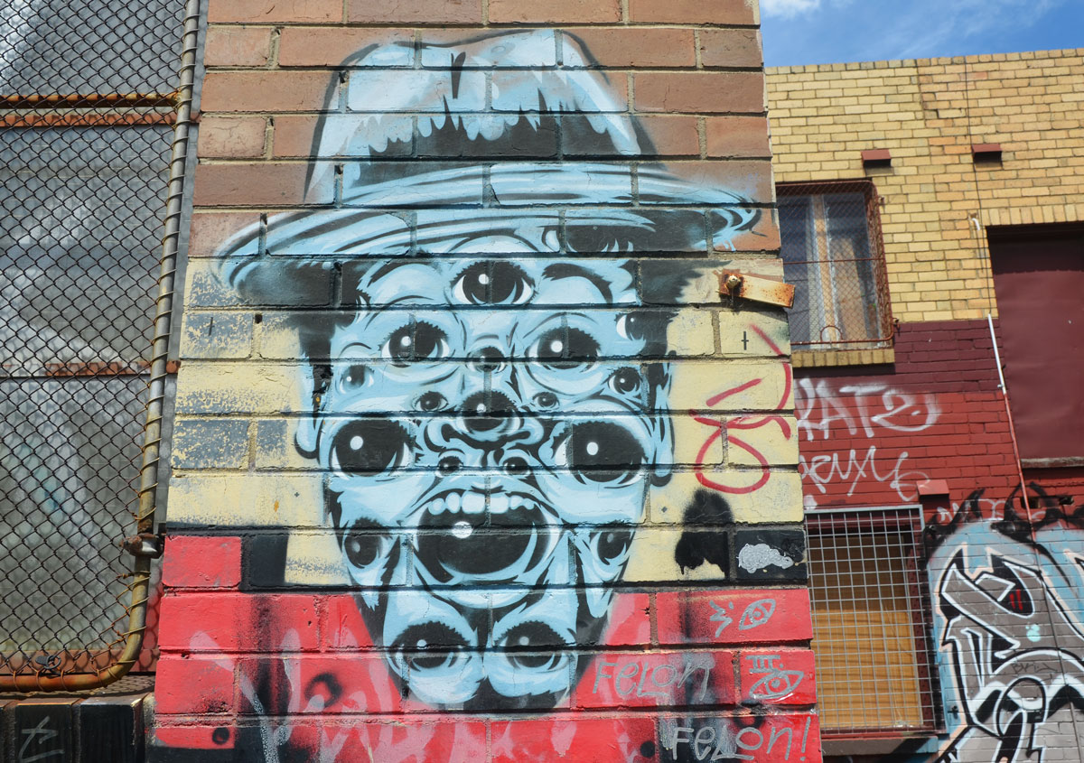 The likeness of a man wearing a hat. All in blue with black details, A mouth, a nose, and many many eyes, on a brick wall, street art 