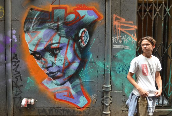 a realistic looking street art painting of a young woman's face in blues. A young man is standing beside painting, in a Melbourne alley