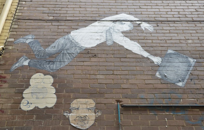 three wheatpaste graffiti pieces on a brick wall . On top is the largest, a man wearing a tie and holding a briefcase is diving horizontally, bleow him are two faces (both wheatpastes)