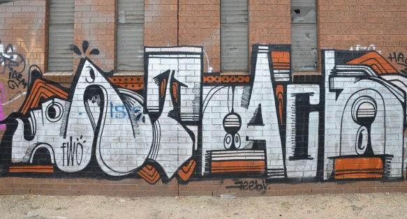 geometric tag in white letters with black and orange accents on a red brick wall. Small letters of the words fwo, feeby and idem