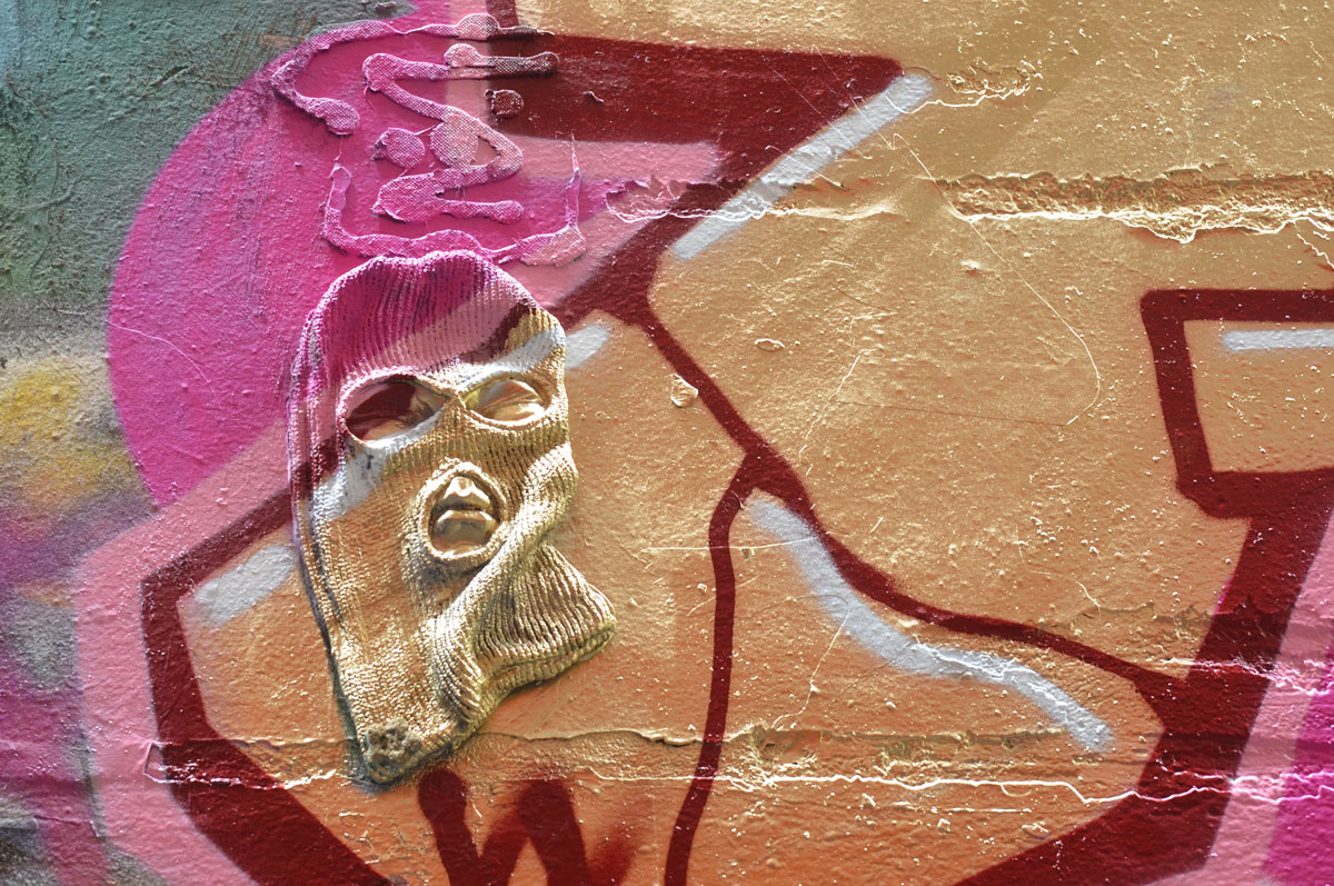 a balaclava mounted on a wall in an alley in such a way that it looks like a face, covered in pink and gold spray paint as part of a street art piece