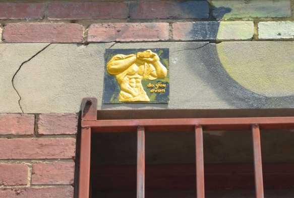 A small relief sculpture of a man's torso painted yellow, headless, large muscles, with hands clasped together in front of his chest holding onto a camera. The words Do you even are written under one elbow.
