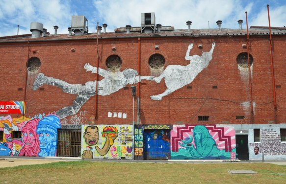wall of an old red brick industrial building, 3 storeys high, covered with street art. Lower level is series of murals and above them is a large paste up by baby guerilla of a couple flying. A man is behind a woman and he is reaching for her foot as she is reaching for the roof of the building.