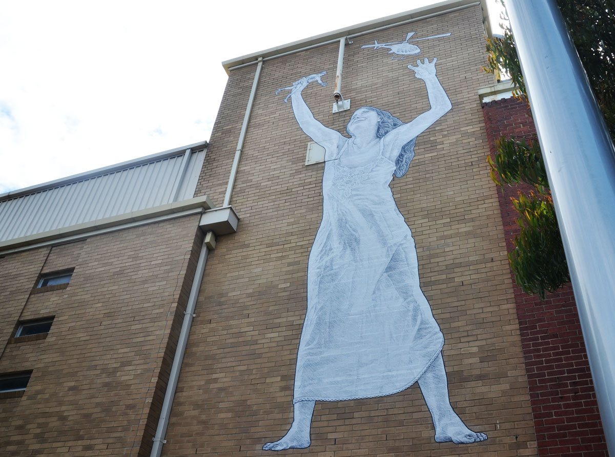 a large paste up of a woman in a long dress holding her hands in the air. In one hand is a helicopter and in the other hand is a small soldier. She is pasted high on a wall and she is about 10 or 12 feet high. 