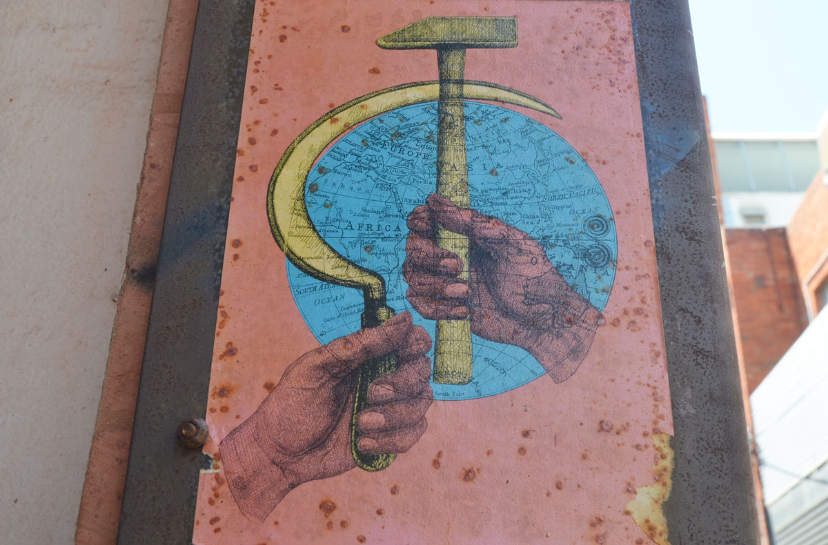 Paste-up of two pink hands, one holding a large hammer and one holding a large sickle. A blue globe (map of the world) is behind the hammer and sickle