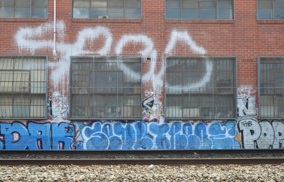 large red brick industrial building beside the train tracks, large windows consisting of many small panes of glass, around the windows there is a lot of graffiti