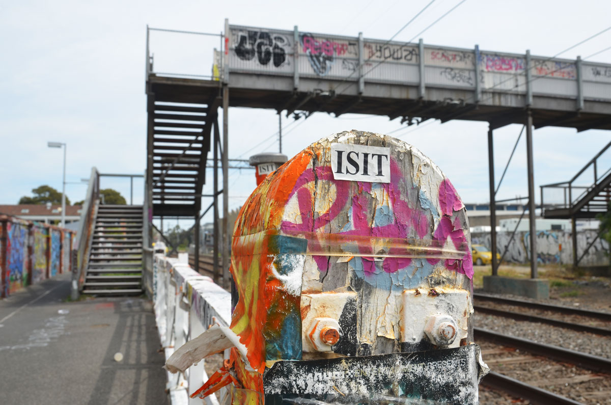 stickers covering a fence post, also spray paint in orange and pink. A white rectangular sticker with ISIT printed on it. In the background, slightly out of focus, is a pedestrian bridge over railway tracks