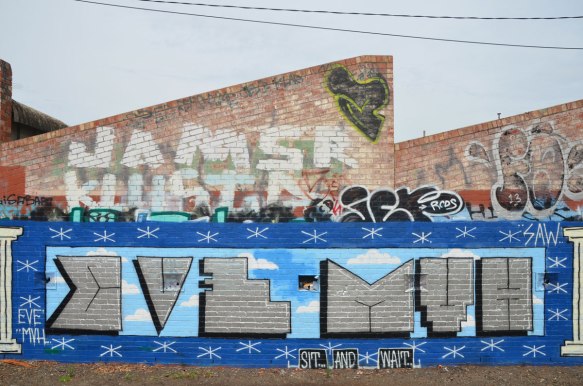 row of old brick buildings with diagonal rooflines covered with graffiti beside a bike path near Jewell train station - large text graffiti that says Eve MVH and the words "sit and wait" written underneath. Jamsr Klustr written in large letters above it. 